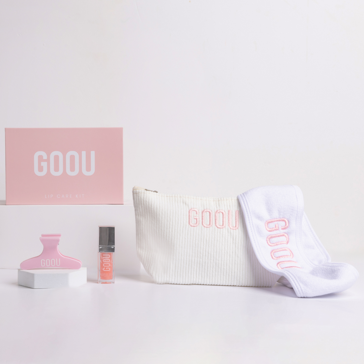 Goou Beauty cosmetic makeup beauty travel bag in white with pink logo featuring lip care kit, lip oil gloss, lip scrub, lip balm mask, pink hair clip grip and white spa headband on white background